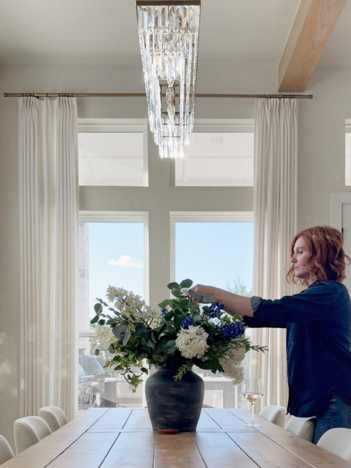 Arranging a fresh floral centerpiece at a light wood dining table beneath a crystal chandelier — a Well Composed Home dining room designed for gathering